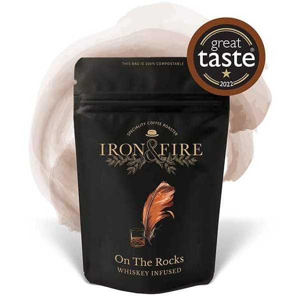 A black coffee bag labeled Iron & Fire On the Rocks Whiskey Infused Speciality Coffee Beans features a copper feather and glass illustration. With Espresso-like depth, it showcases a Great Taste 2022 award badge against a subtle light brown artistic splash backdrop.