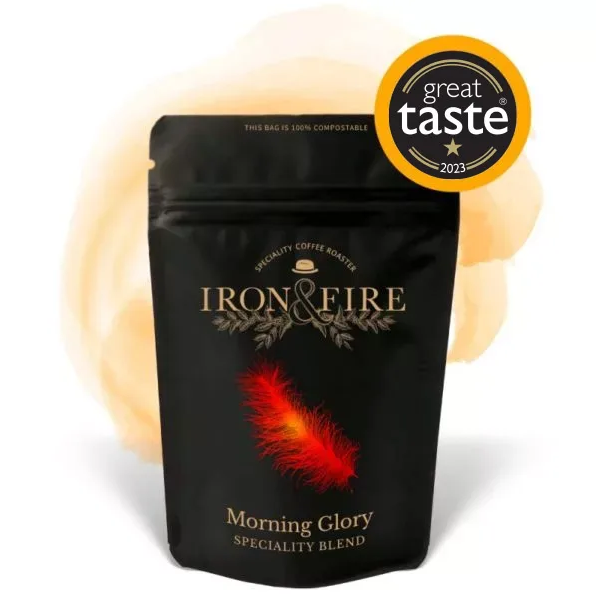 The Iron & Fire Morning Glory Speciality Coffee Beans come in a black bag with a red feather design, boasting an award-winning speciality blend, zesty orange flavors, and a Great Taste 2023 badge. The 100% compostable packaging enhances the appeal of its single-origin beans.