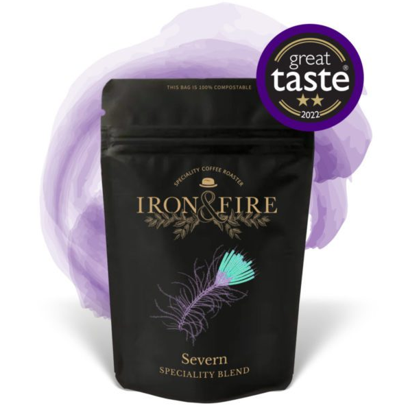 The Iron & Fire Severn Blend Coffee Beans, in a black bag with a feather design and Great Taste 2022 award sticker, stands against a purple watercolor background. It promises full-bodied coffee with rich chocolate flavors and comes in a 100% compostable package.