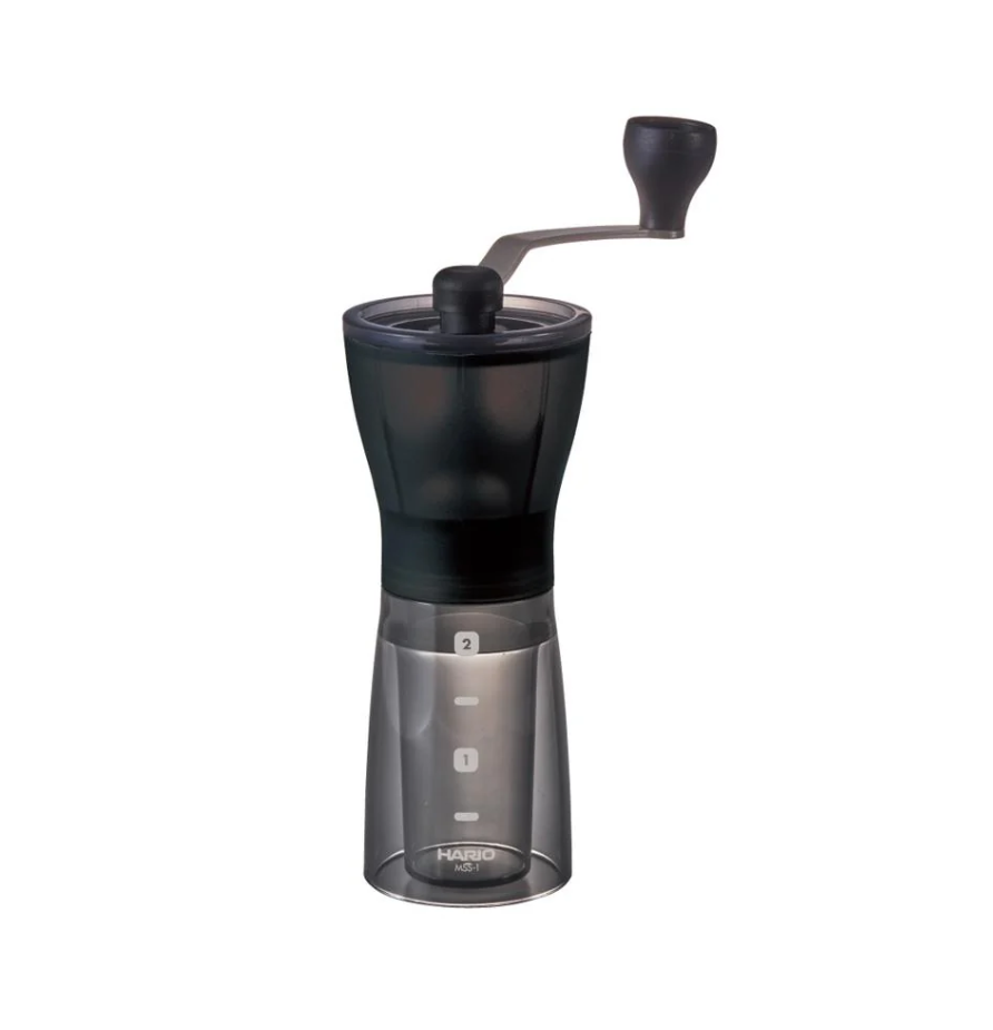 The Hario Mini Mill Plus is a ceramic coffee grinder with a black upper body, transparent lower chamber, hand crank on top, measurement markings, and the Hario brand name at its base.