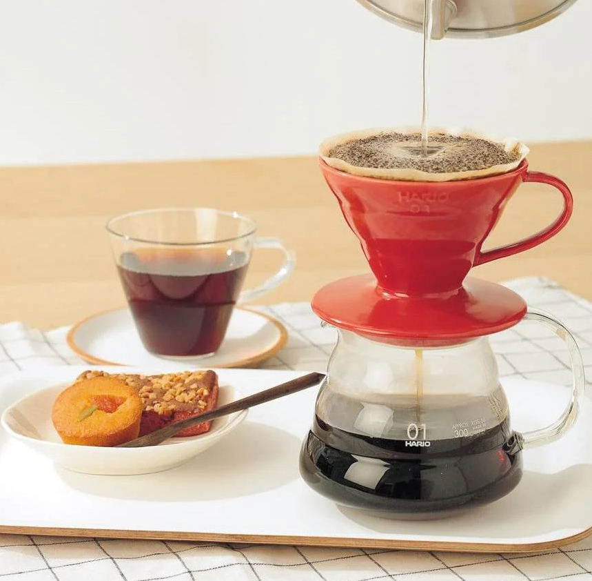 A Hario V60 Ceramic Coffee Dripper - Red (Size 01) by Hario is used for a pour-over on a glass carafe, with coffee served beside cake and a muffin, while the kettle pours water over the grounds in this cozy setup featuring quality coffee equipment.