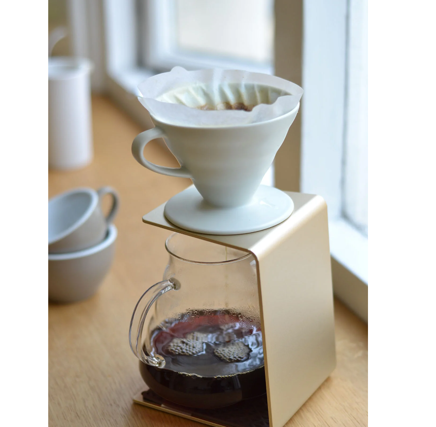 A red Hario V60 Ceramic Coffee Dripper sits with a filter on a glass carafe filled with freshly brewed coffee on a wooden table. Nearby, two empty cups are illuminated by natural light from the window, showcasing this exquisite piece of Hario coffee equipment.