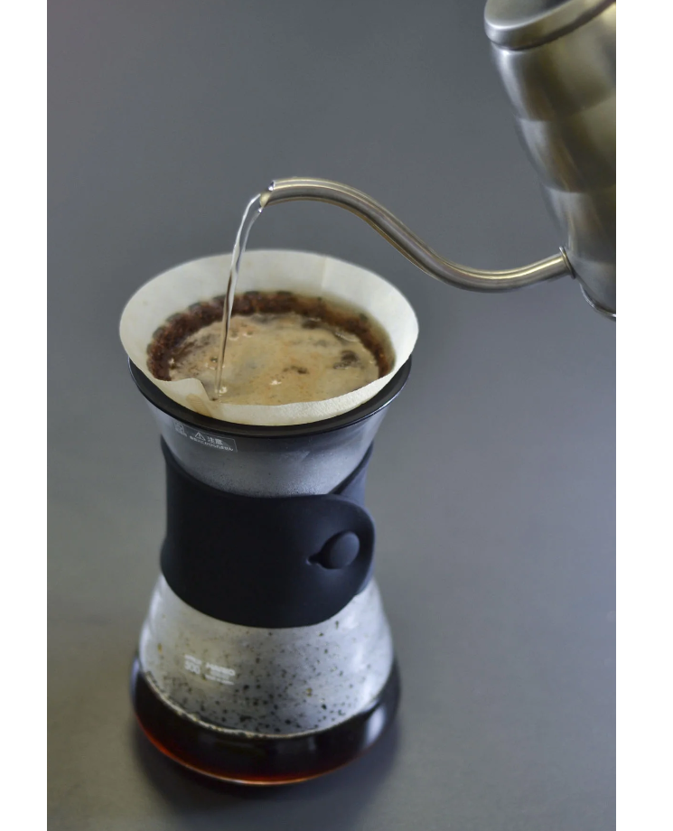 A Hario V60 Drip Decanter Pour Over Coffee Maker pours hot water over a coffee filter in a ceramic Chemex to brew coffee, set against a simple gray background.