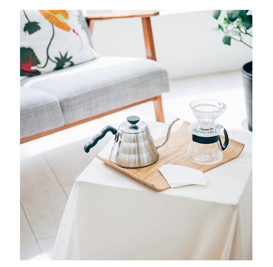 A cozy setup includes a wooden tray with a stainless steel gooseneck kettle and glass pour-over dripper. Nearby, an armchair with a floral cushion is visible alongside a plant, enhancing the homey feel with the charm of a Hario V60 Glass Coffee Server - Size 01 (360ml) by Hario.