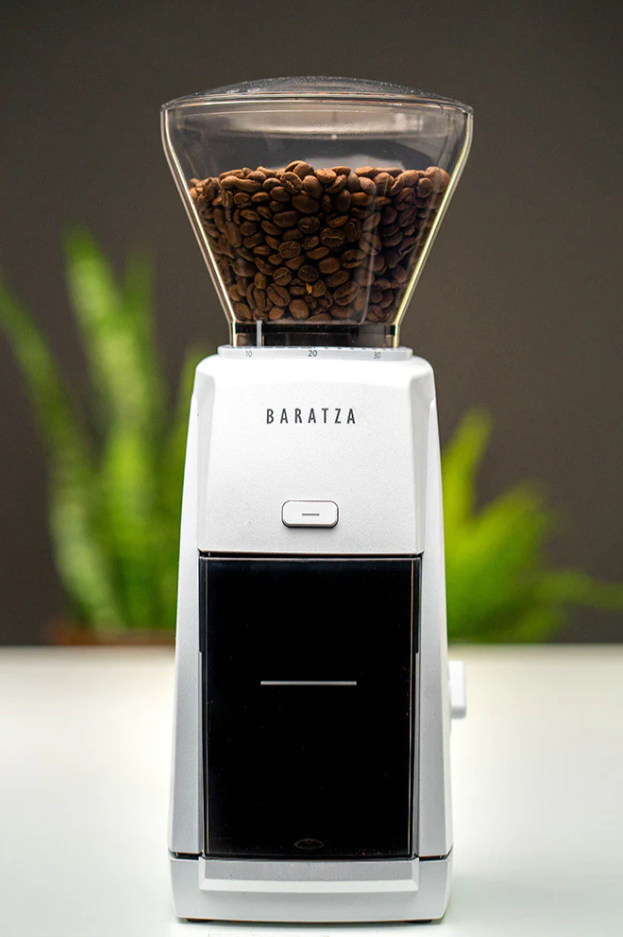 A white Baratza Encore ESP Coffee Grinder, optimized for espresso and filled with specialty beans, sits on a light surface. In the background, a blurred green plant contrasts with a dark backdrop, highlighting its high-resolution grinding capabilities.