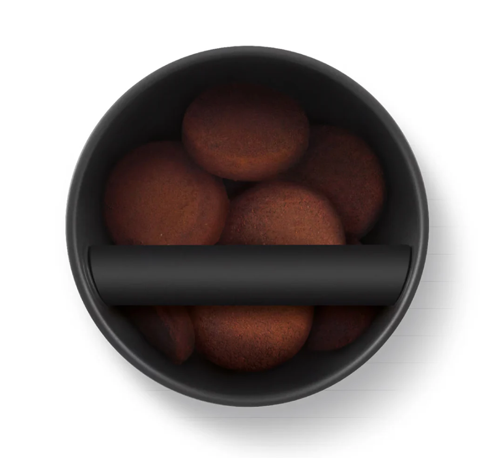 A top-down view of a black Sage The Knockbox Puck Disposal Box with a removable knock bar, partially covering a black bowl filled with round brown cookies, all set against a white background.