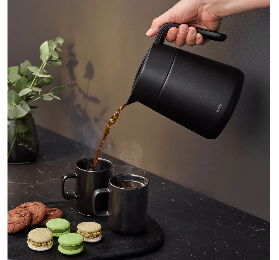 A hand pours coffee from a Wilfa Performance Thermo Coffee Maker carafe into two matching mugs on a black surface with green macarons, cookies, and a vase—highlighting Wilfa’s sleek Scandinavian design.