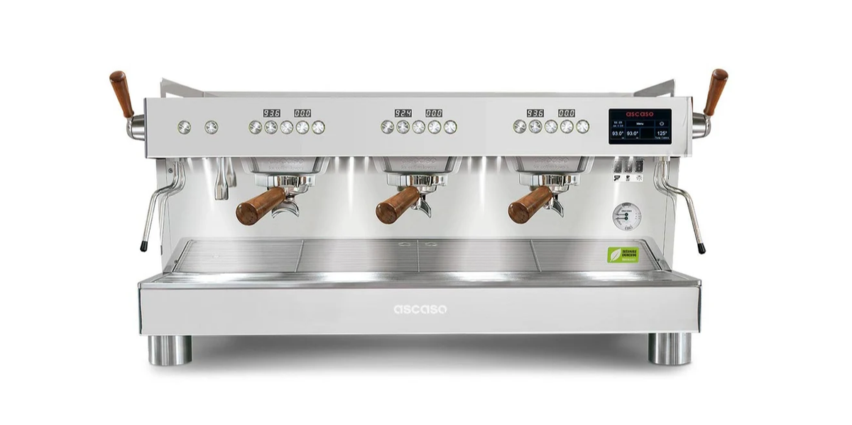 The Ascaso Barista T One 3 Group by Totally Delicious Coffee is a white, professional espresso machine featuring three groups, elegant wooden handles, a stainless steel drip tray, digital display, and multiple control buttons. Shown on a white background, this energy-efficient model blends sleek style with modern functionality.