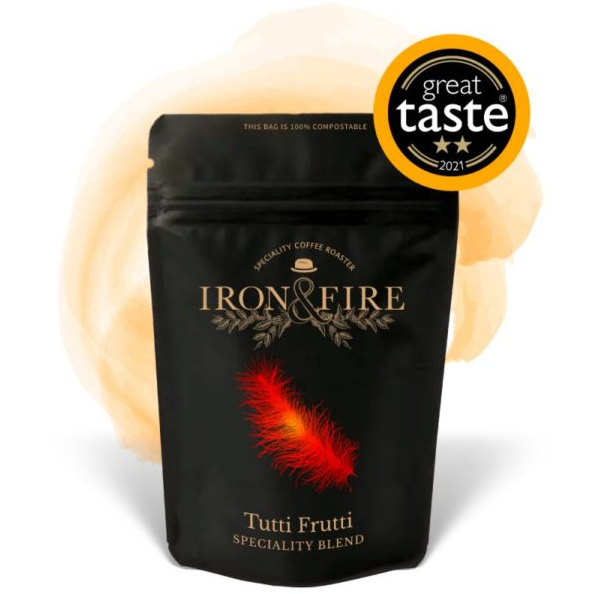 The Iron & Fire Tutti Frutti Speciality Coffee Beans is presented in a black bag featuring a feather design and a Great Taste 2021 award sticker, showcasing its UTZ certification and floral notes against an orange watercolor splash background.