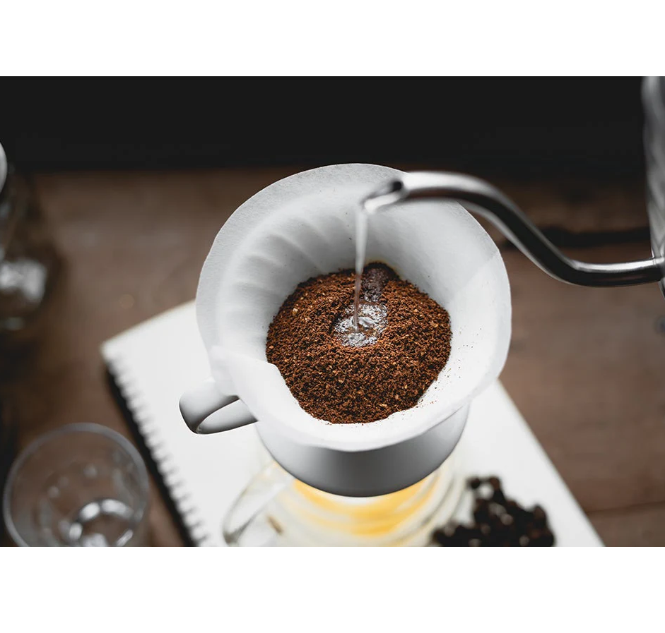 An overhead view of a Hario V60 pour-over setup shows a kettle pouring hot water on coffee grounds in Size 02 Coffee Filter Papers within the dripper as brewed coffee flows into the glass carafe. A notebook and glass appear in the background.