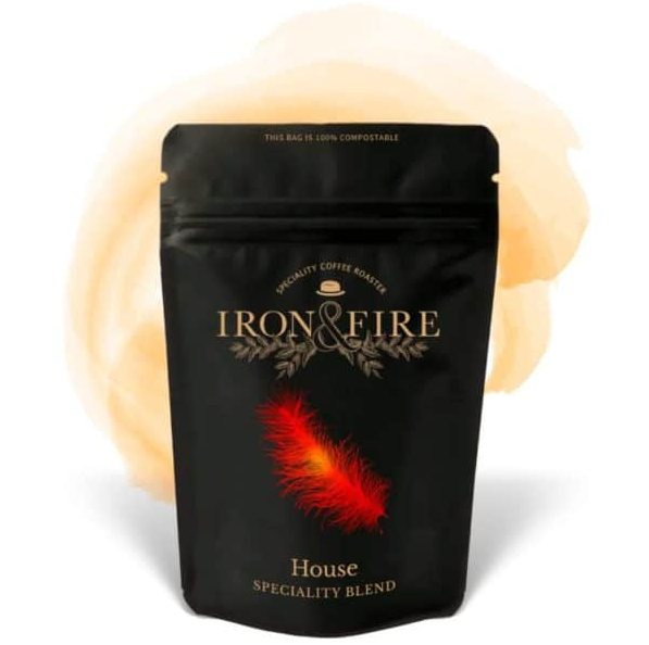 The Iron & Fire House Speciality Blend Coffee Beans come in a black bag featuring gold text and a decorative feather design. Below, House Speciality Blend is highlighted, set against a soft orange watercolor splash. The bag, made from smallholder-sourced Colombian and Brazilian beans, is 100% compostable.