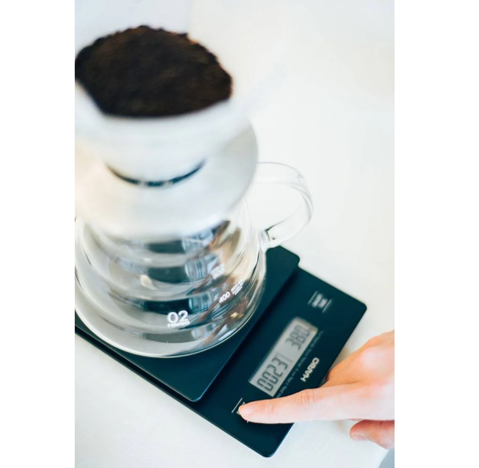 A person adjusts a Hario V60 Drip Coffee Scale - Black beneath a glass dripper loaded with ground coffee. The scales display shines, enhancing the bright and minimalistic scene with its softly blurred background.